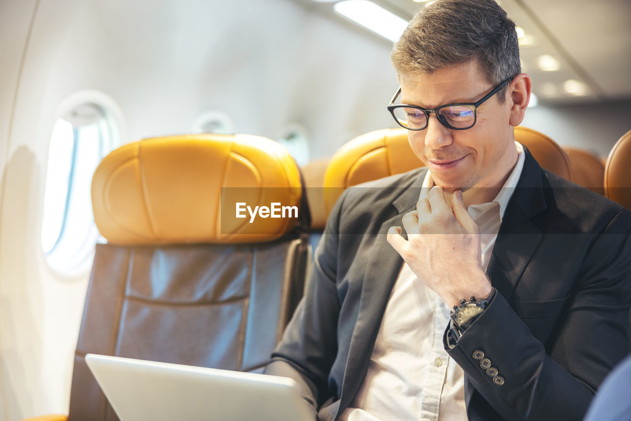 During a flight, a businessman in formal attire and glasses looks out the plane window while working