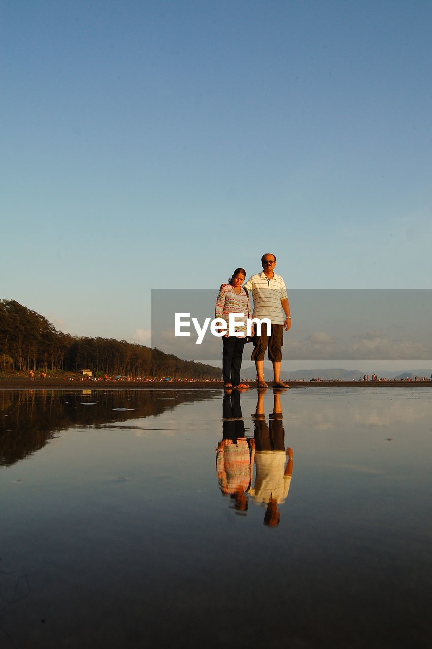 Couple standing at lakeshore against sky during sunset