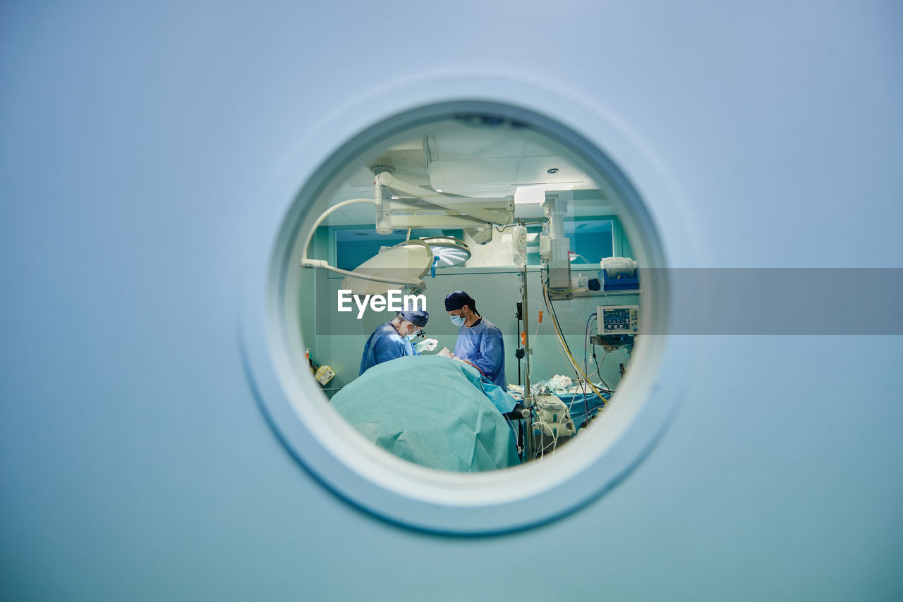 Through round window of unrecognizable doctors in uniforms and masks performing surgery in modern operating room