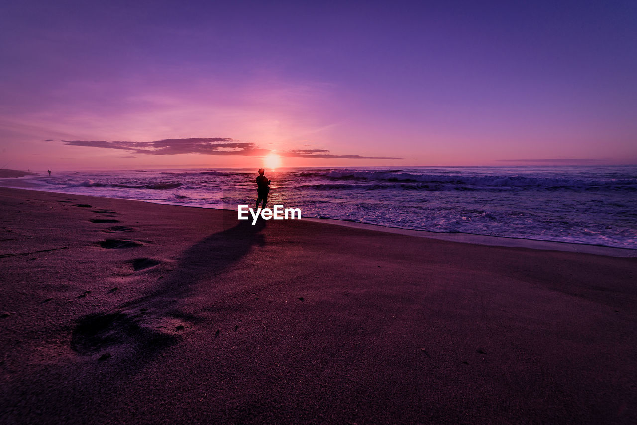 Silhouette man standing on shore at beach against purple sky during sunset