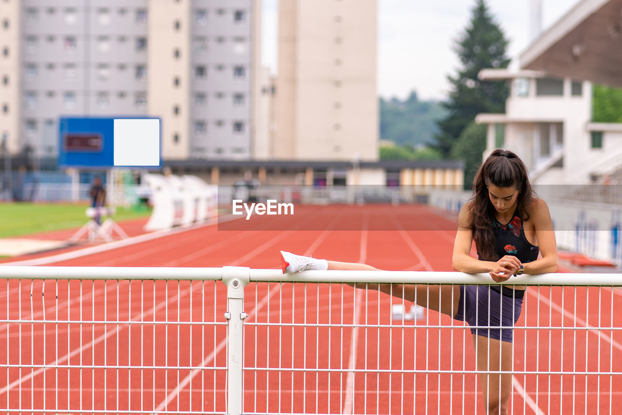 Young hispanic female athlete stretching legs near metal railing while warming up before running on stadium track