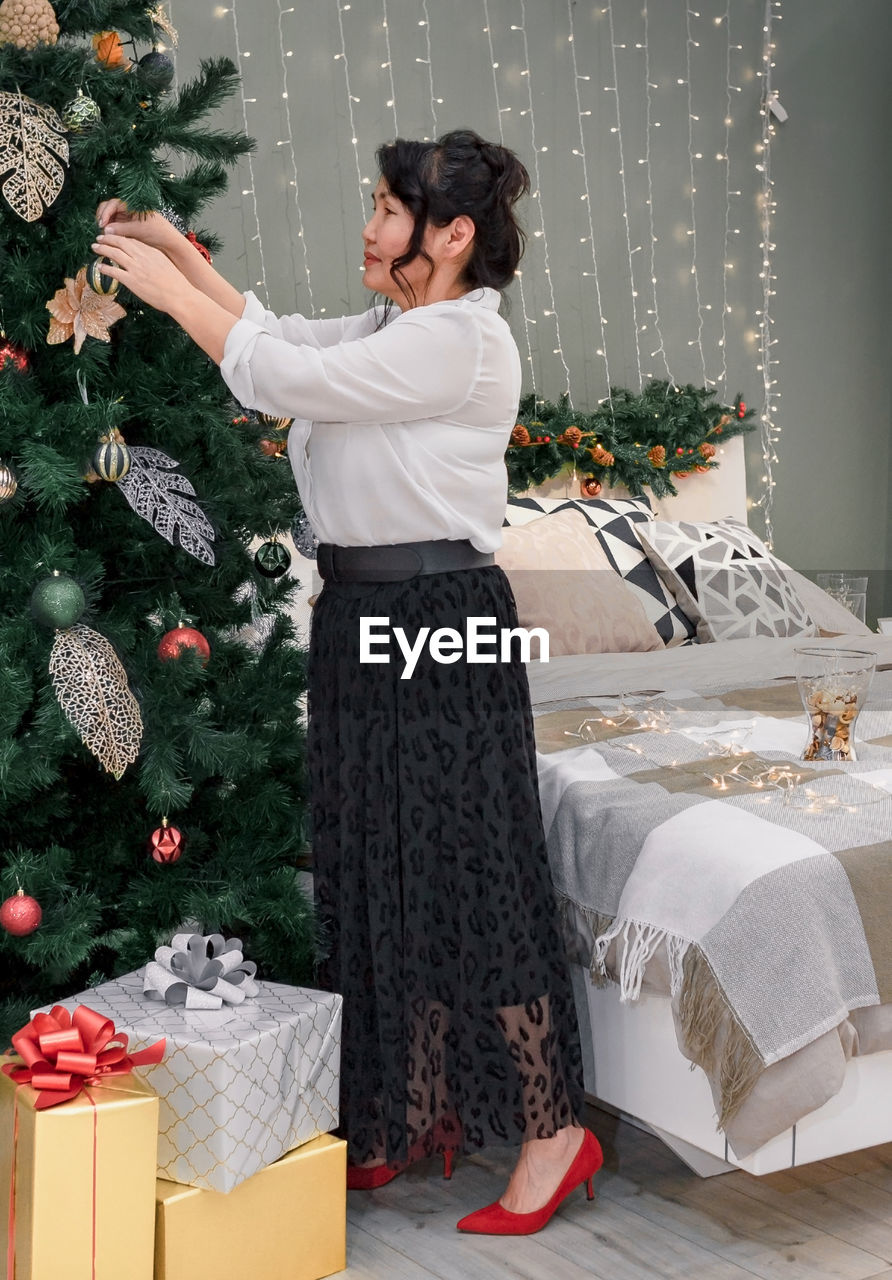 Senior asian woman decorate christmas tree by xmas bauble decoration in bed room with gift at home