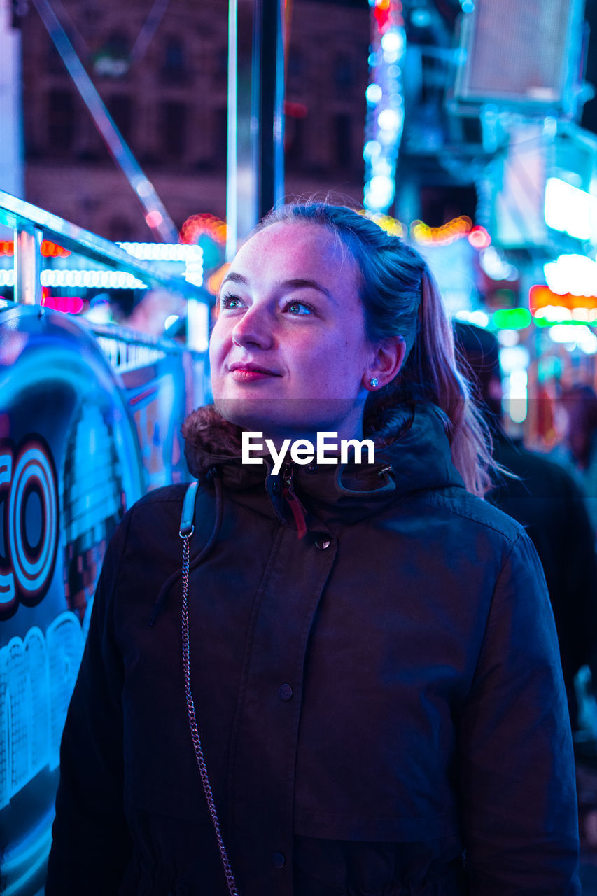 Close-up of young woman standing against illuminated amusement ride at night