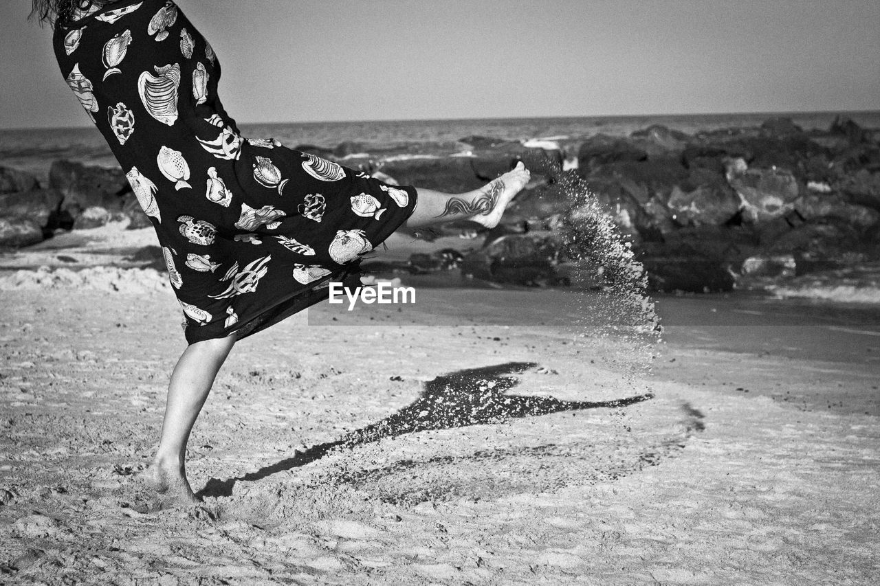 Low section of woman kicking sand at beach against sky