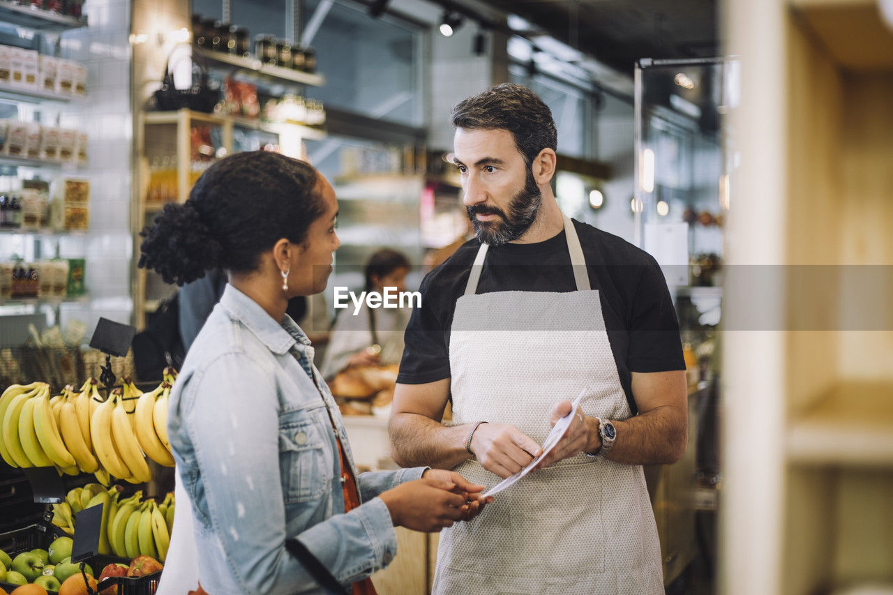 Mature salesman discussing over document with female customer at delicatessen