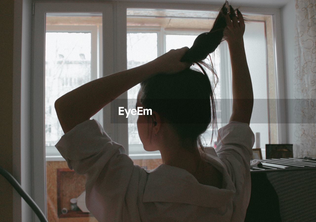 Woman tying hair while sitting against window at home