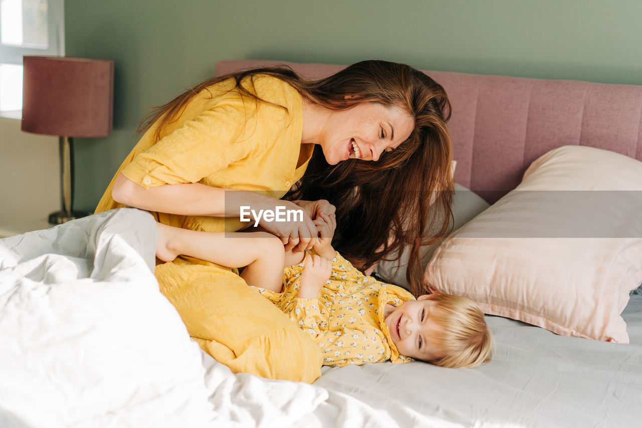 Young red-haired mother plays and tickles her little cheerful daughter sitting on the bed.