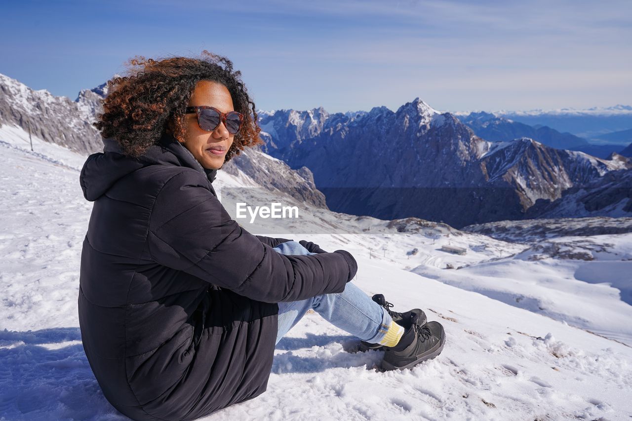 Pretty young woman is sitting in the snow, looking at the mountain view/ skiing area/ zugspitze