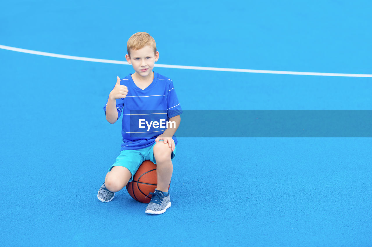 Portrait of boy gesturing thumbs up while sitting on basketball at court