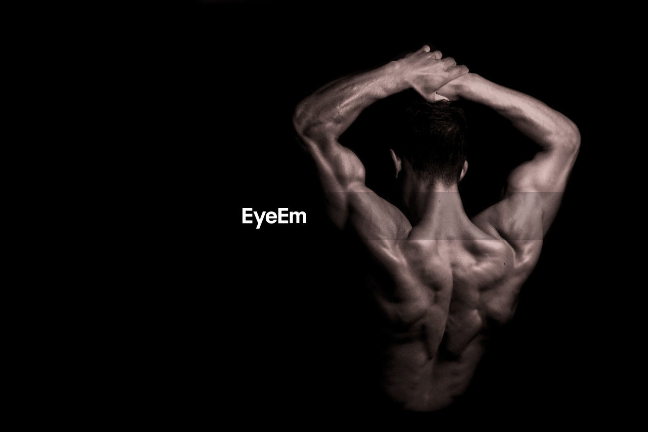 Rear view of shirtless body builder with arms raised against black background