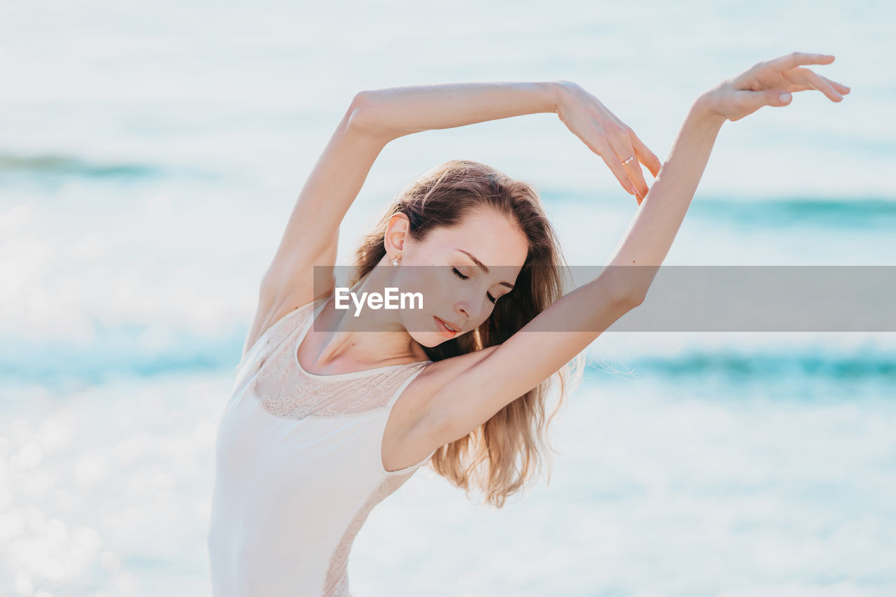 Beautiful young woman dancing with eyes closed at beach against sea