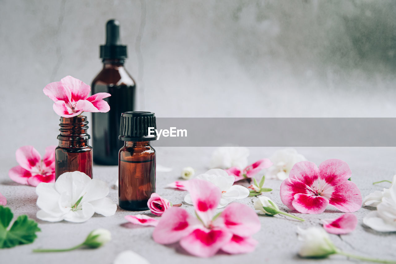 Front view of glass bottles of geranium essential oil with fresh pink and white flowers and petals.