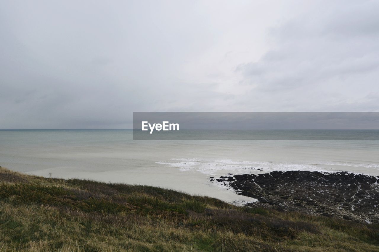 sea, water, land, sky, beach, horizon, coast, shore, beauty in nature, scenics - nature, ocean, cloud, body of water, horizon over water, nature, wave, tranquility, environment, cliff, landscape, tranquil scene, coastline, no people, grass, rock, bay, wind wave, terrain, day, seascape, non-urban scene, outdoors, overcast, plant, travel destinations, sand, storm, travel, idyllic, tourism, dramatic sky, water's edge, motion