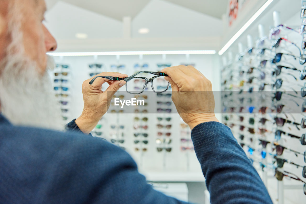 Serious elderly bearded male in trendy outfit standing with modern stylish glasses in optical shop