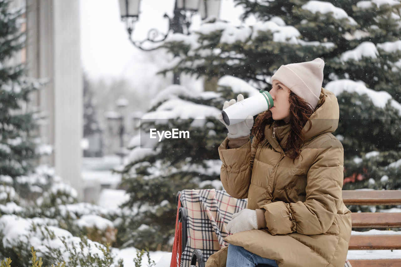 Cheerfully brunette woman drinking from flask outdoors in the city in winter. warming up, enjoying