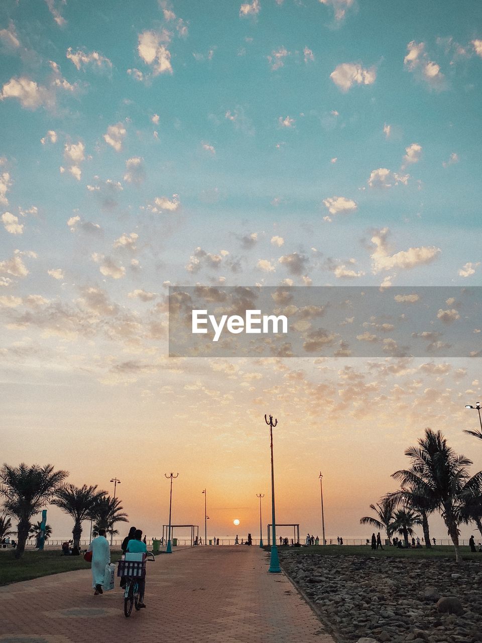 PEOPLE ON ROAD BY PALM TREES AGAINST SKY AT SUNSET