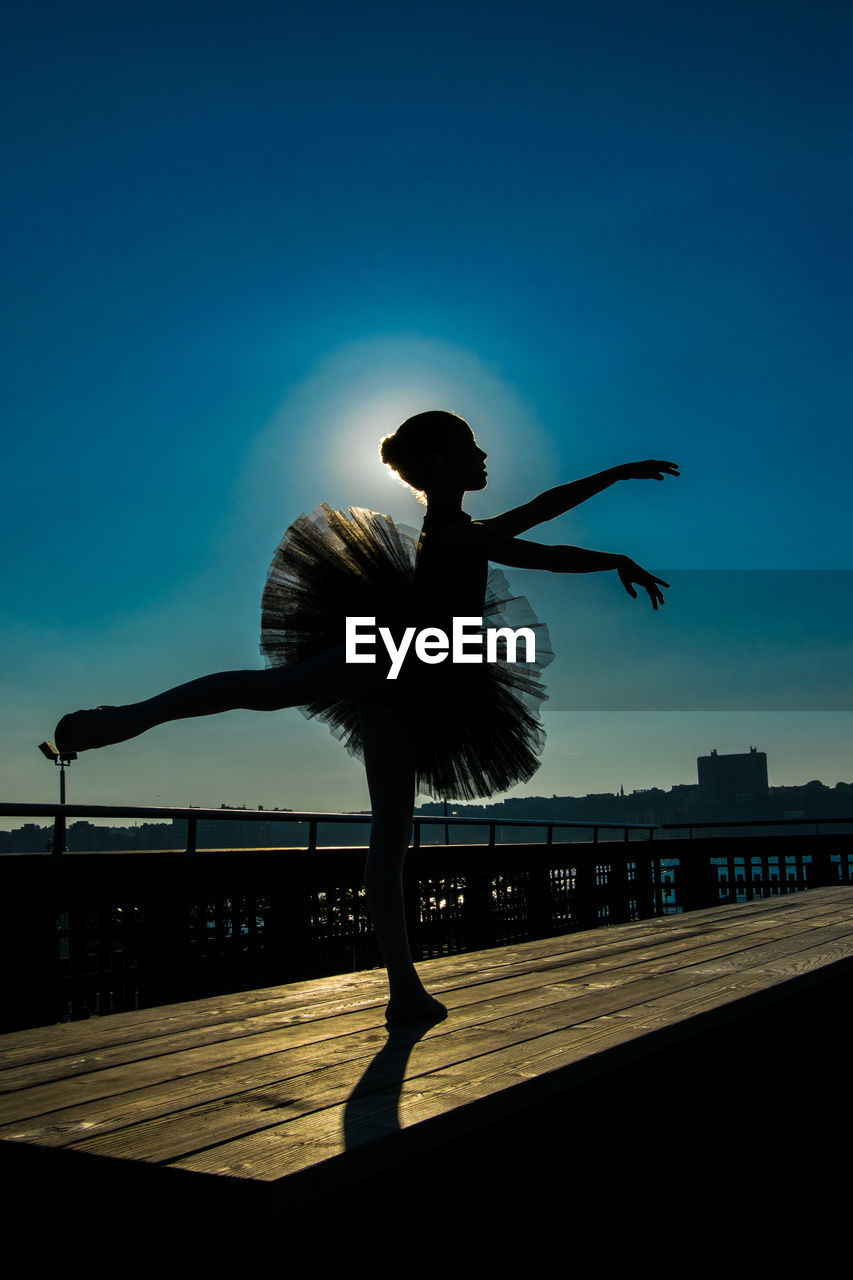 Silhouette ballerina dancing on wooden table at pier against sky during sunny day