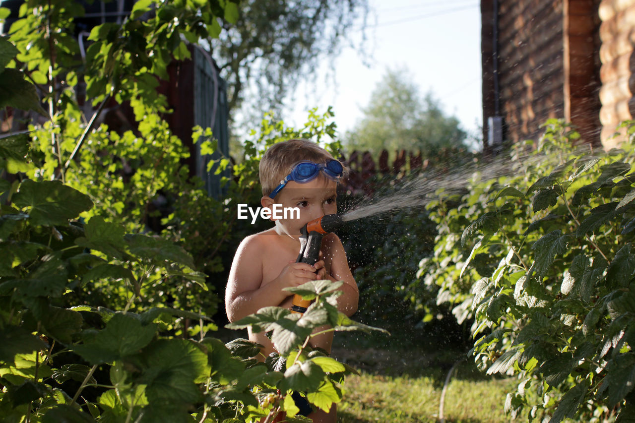 Happy little boy in watersport goggles with a pistol in hands watering the garden in summer cottage