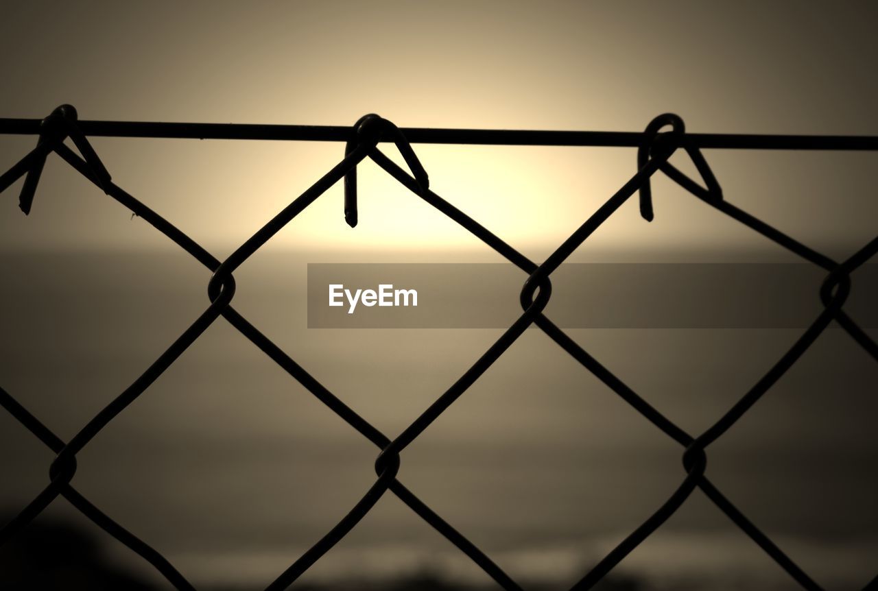 Close-up of chainlink fence against sea during sunset