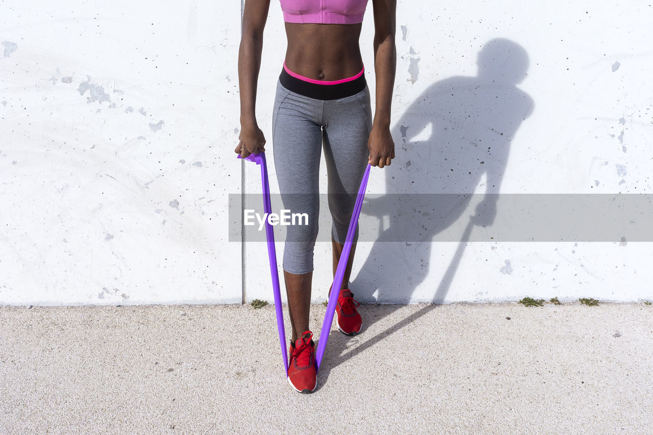 Young young woman doing arm workout using rubber band against white wall during sunny day