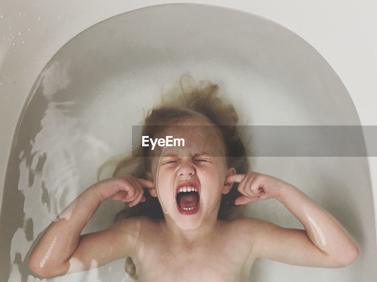 Directly above shot of shouting shirtless girl covering her ears in bathtub