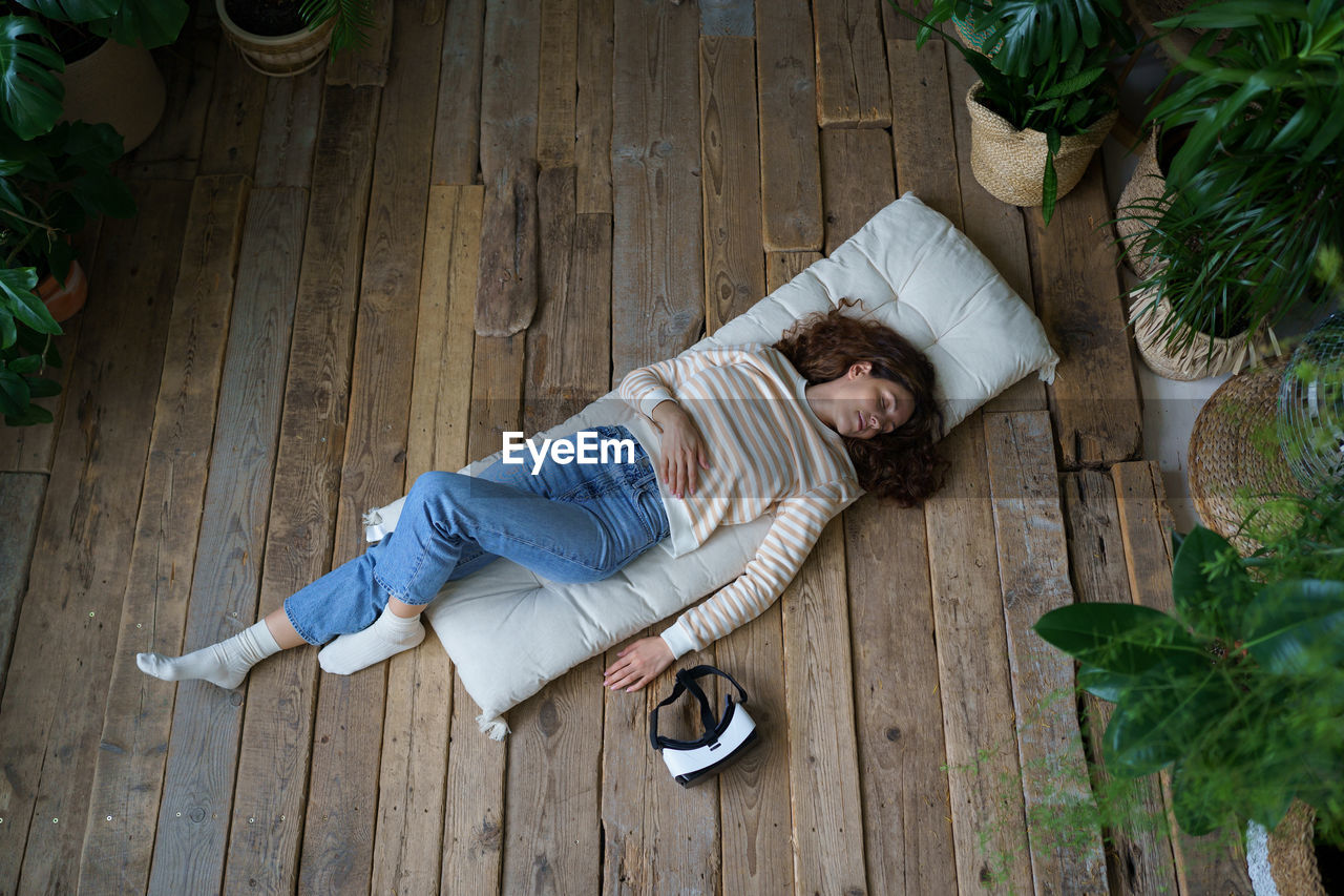 Relaxed woman resting after vr session, sleeping on wooden floor surrounded by tropical plants