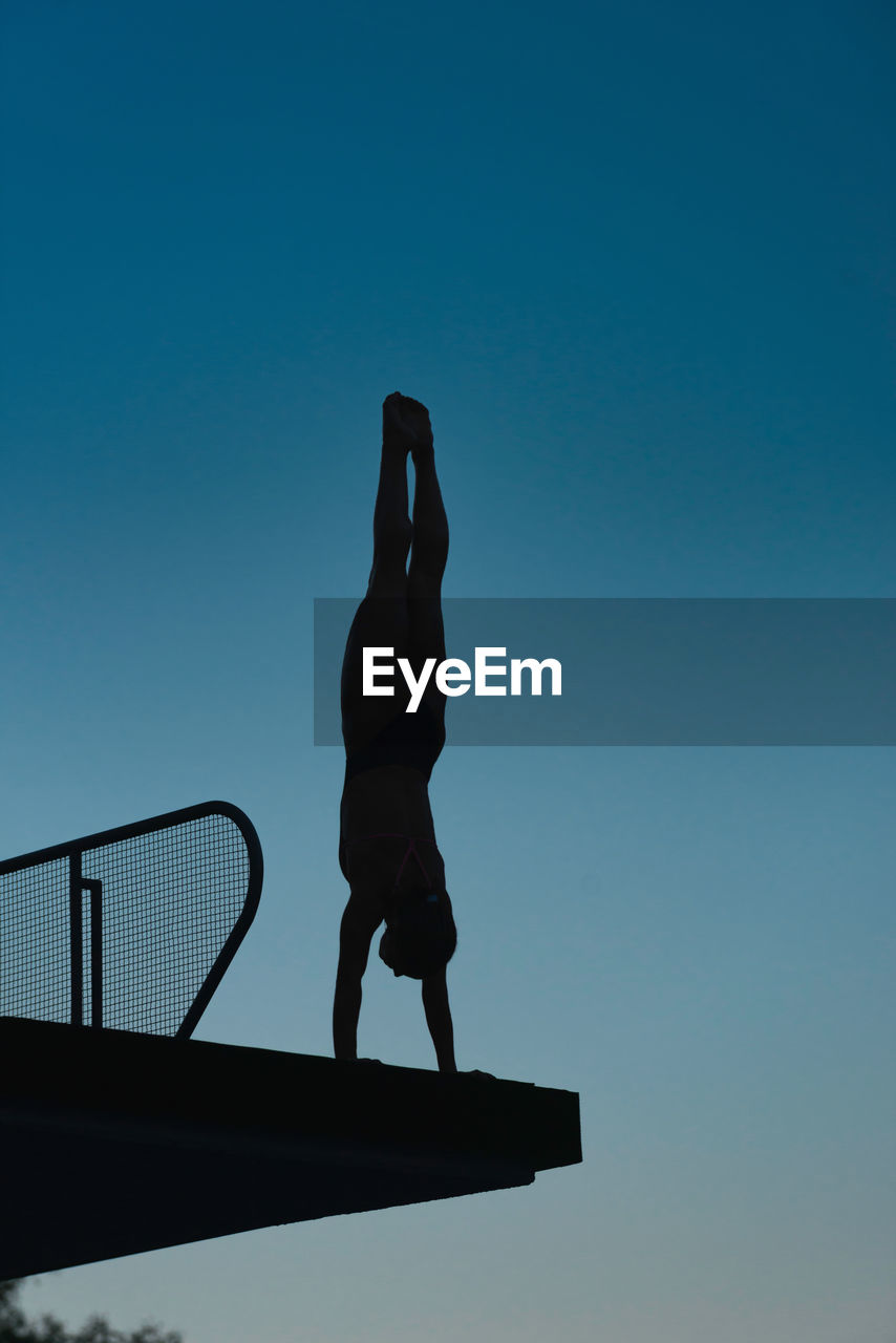 Low angle view of silhouette mid adult woman doing handstand on diving platform against clear sky during sunset