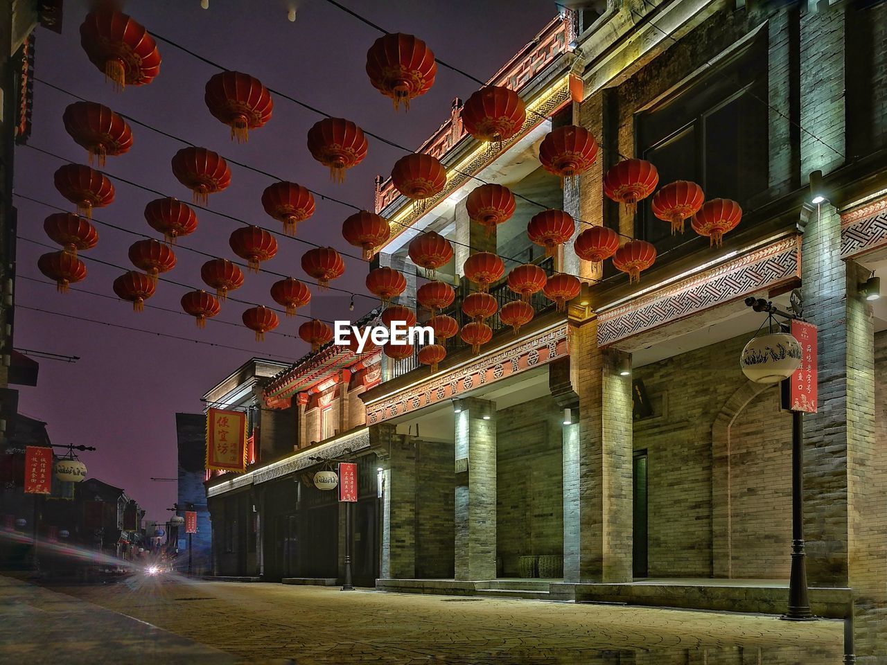 LOW ANGLE VIEW OF ILLUMINATED LANTERNS HANGING BY BUILDINGS AT NIGHT