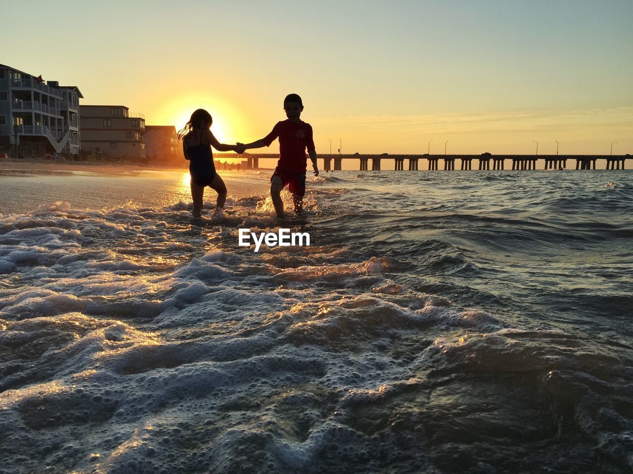 Playful siblings dancing at shore against sky during sunset
