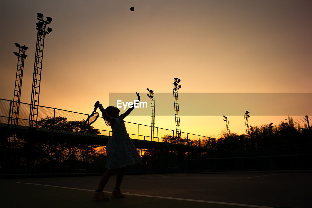 Silhouette girl playing tennis while standing in court against sky during sunset