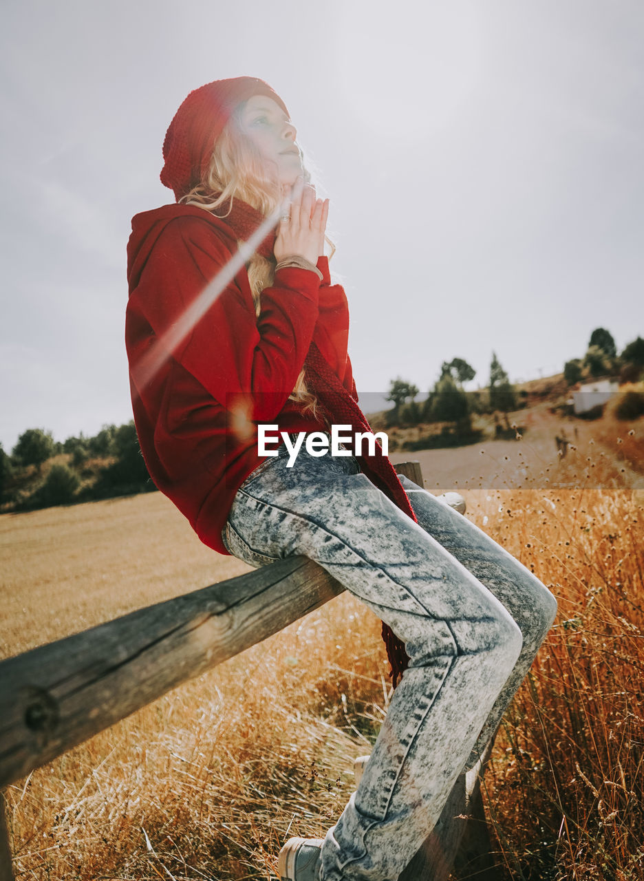 Woman sitting on railing in field against sky