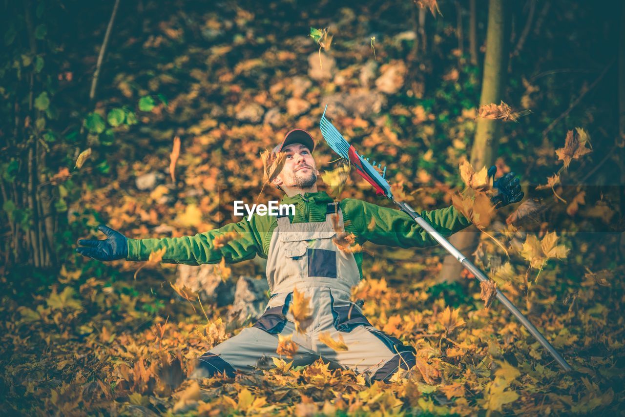 Mid adult man with arms outstretched kneeling on autumn leaves by gardening fork