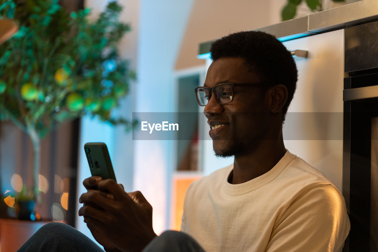 Young happy black guy using smartphone while relaxing and spending leisure time at cozy home