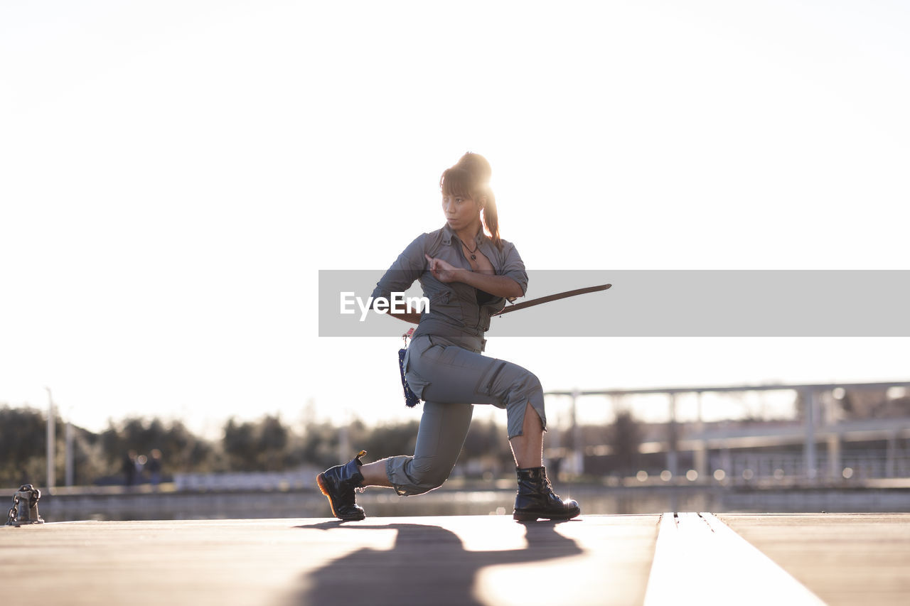 Young female martial artist kneeling with sword at park on sunny day
