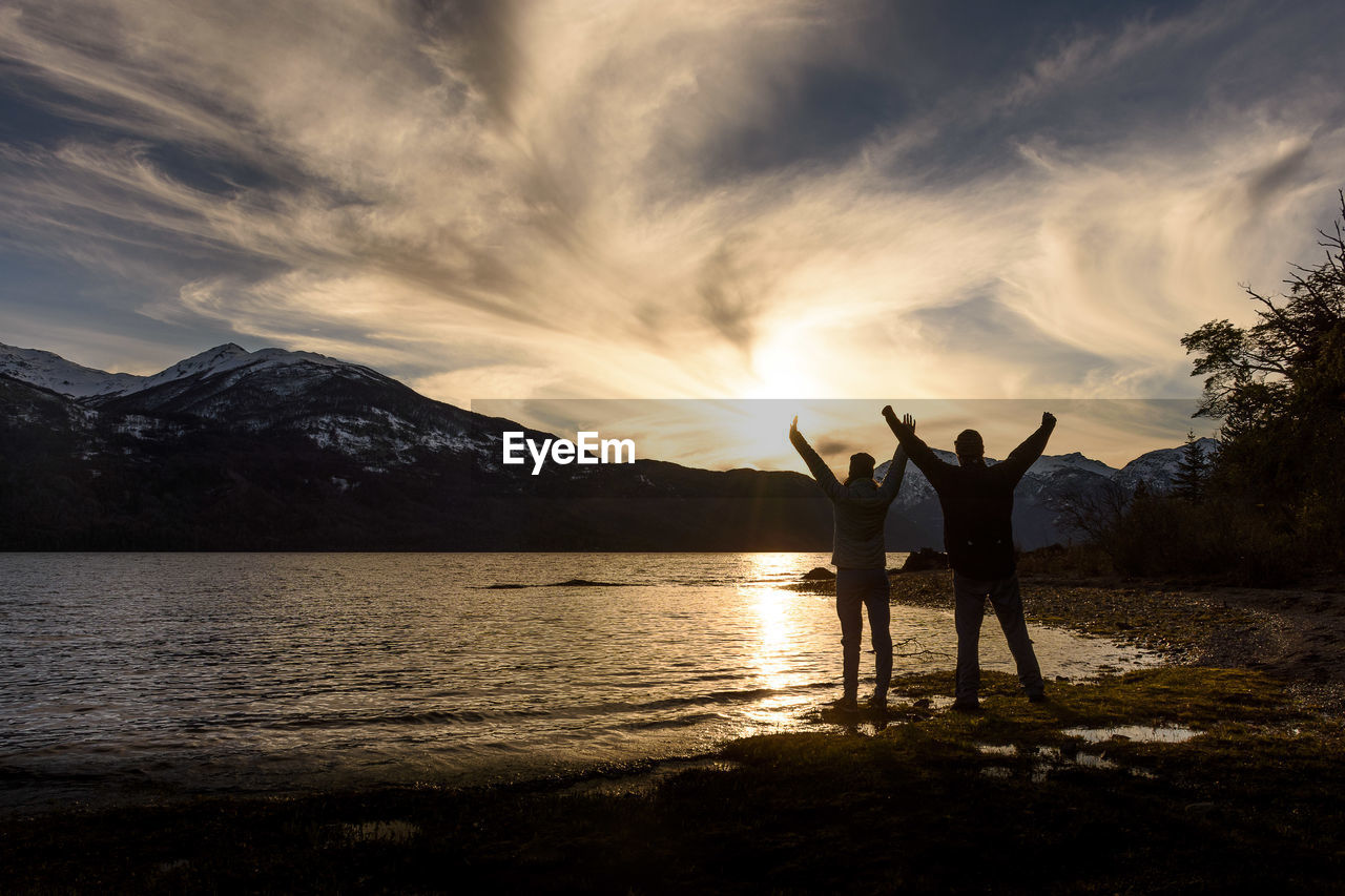 Friends standing by lake against sky during sunset