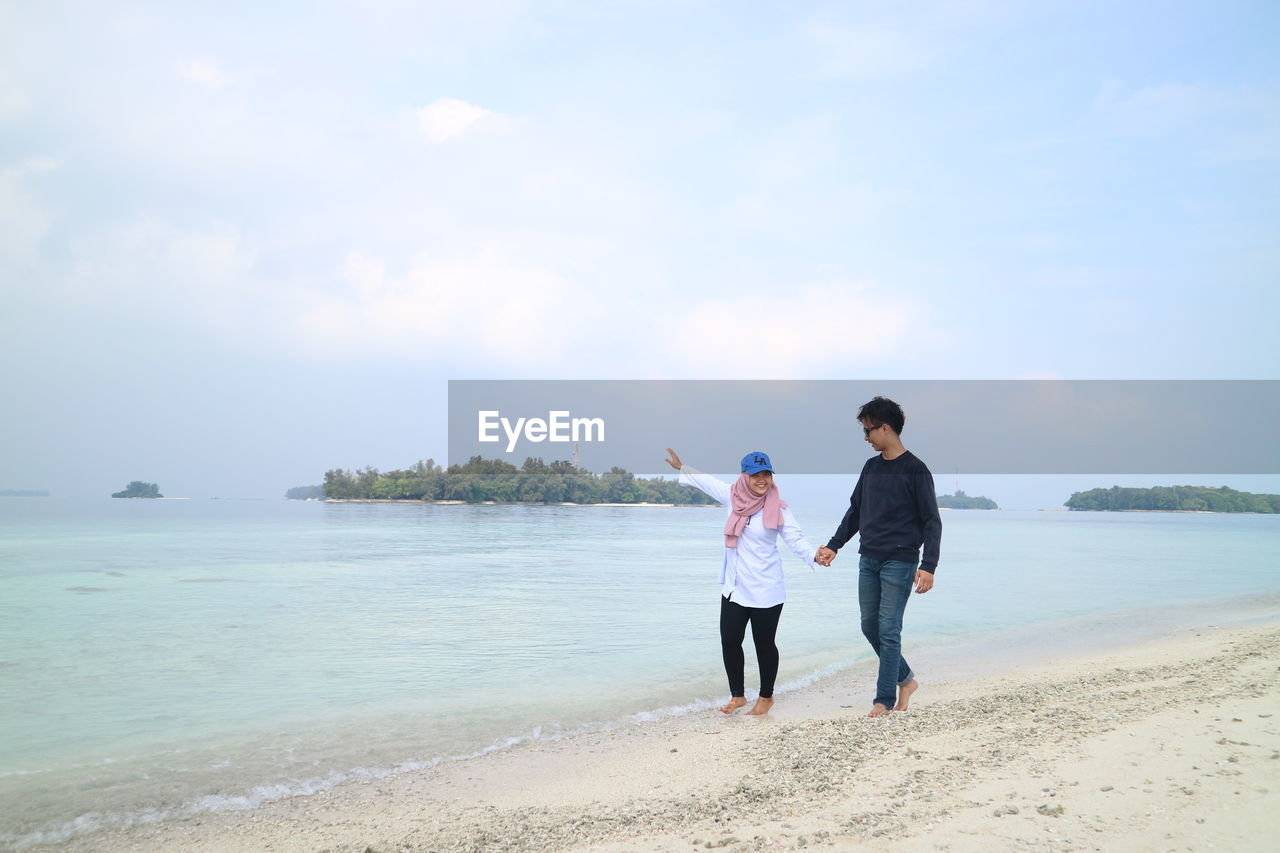 Full length of young couple holding hands while walking on shore at beach against sky