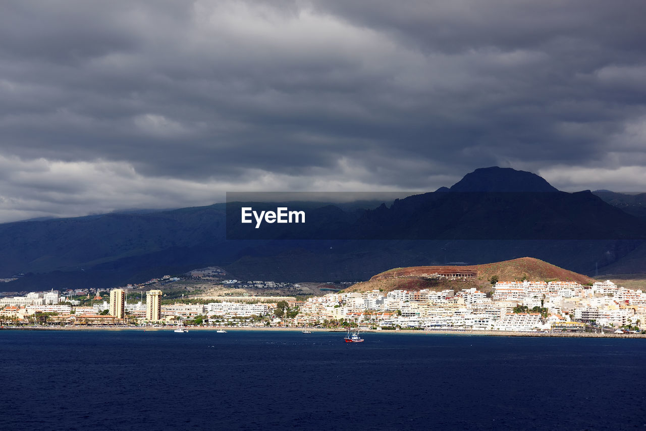 Sea and community by mountains against cloudy sky