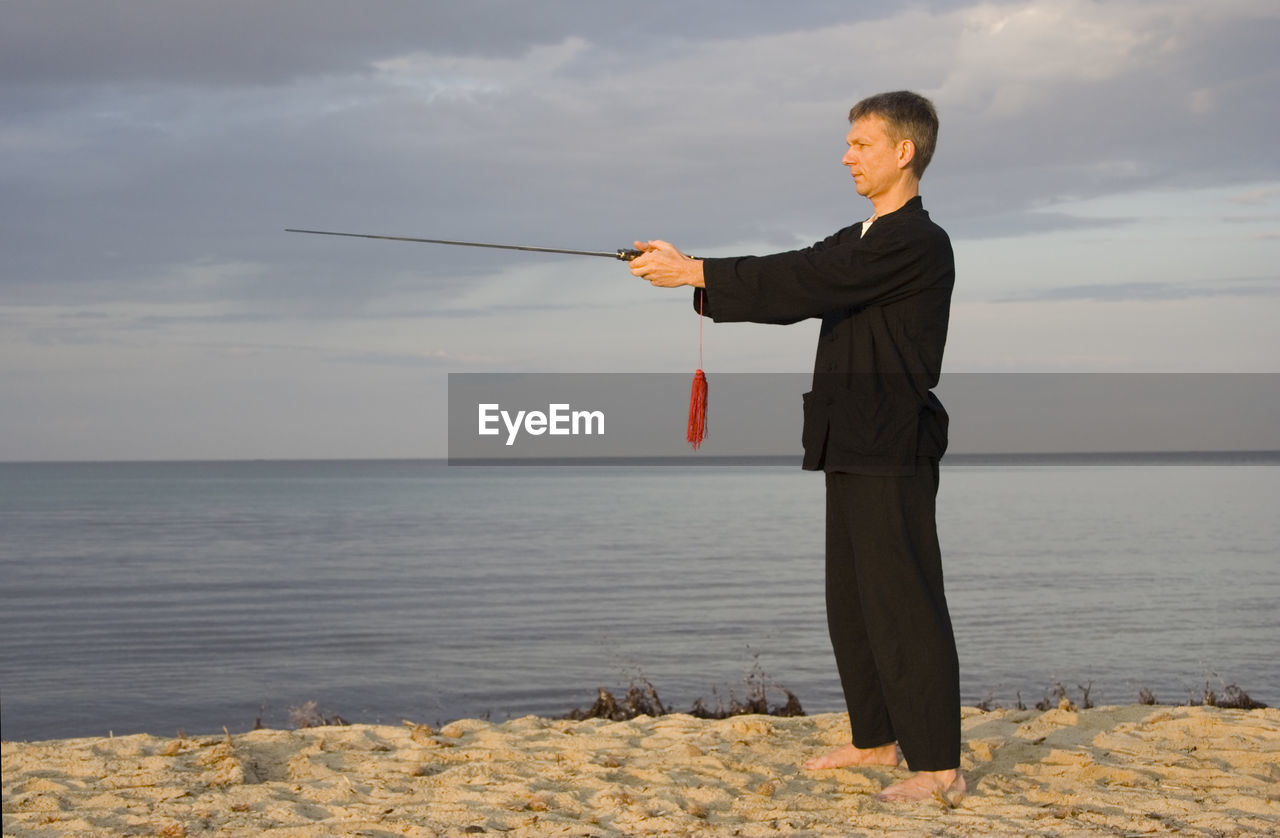 Side view of man practicing martial arts with sword at beach against sky during sunset
