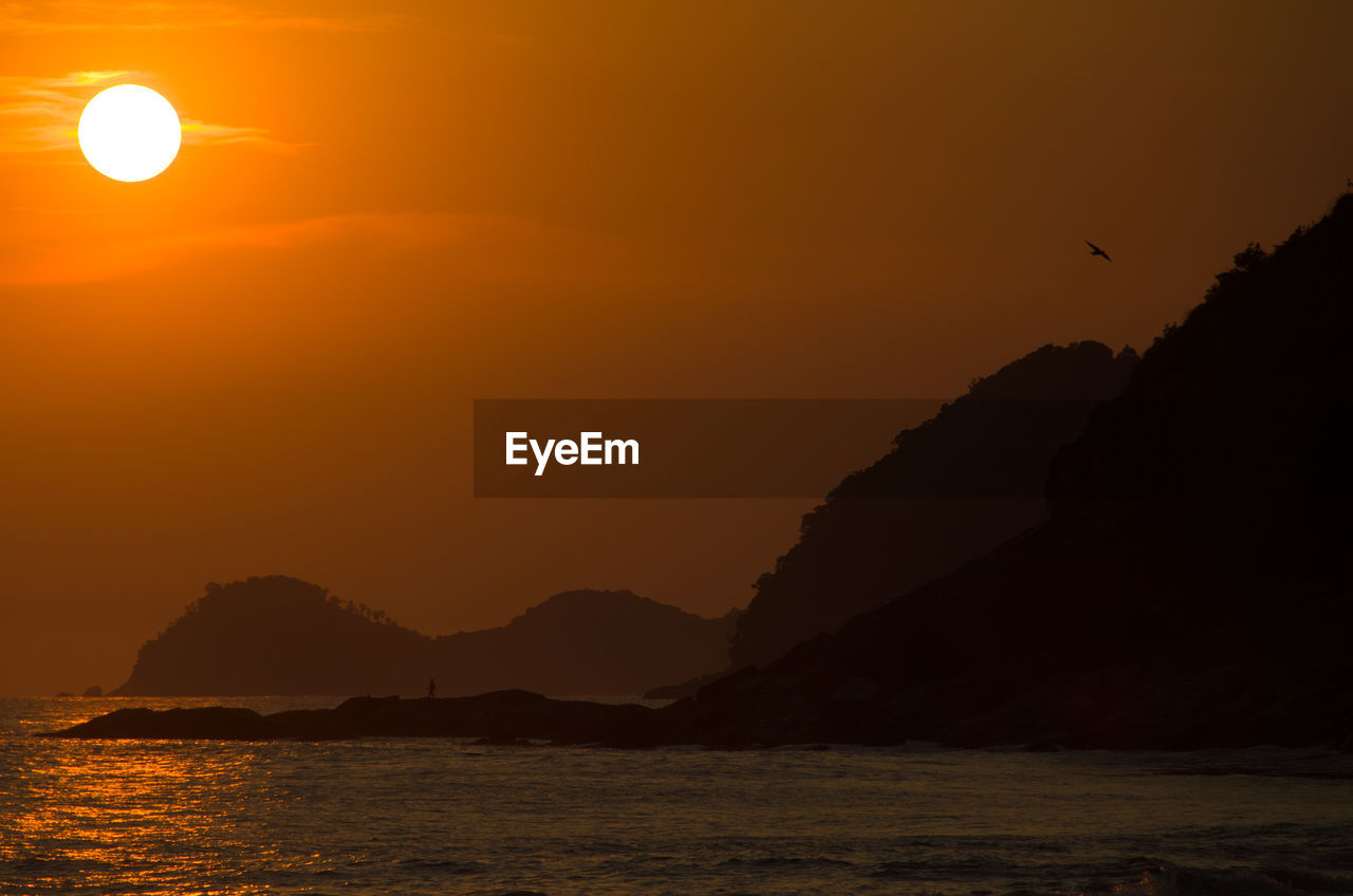 SCENIC VIEW OF SEA AND SILHOUETTE MOUNTAINS AGAINST ORANGE SKY