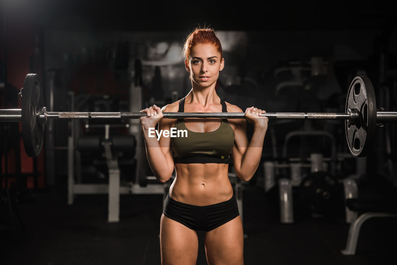 Mid adult woman looking at camera holding barbell olympic bar in gym during intense workout