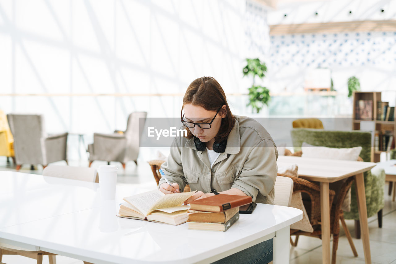 Young teenager girl college student in glasses doing homework with books at green modern library 