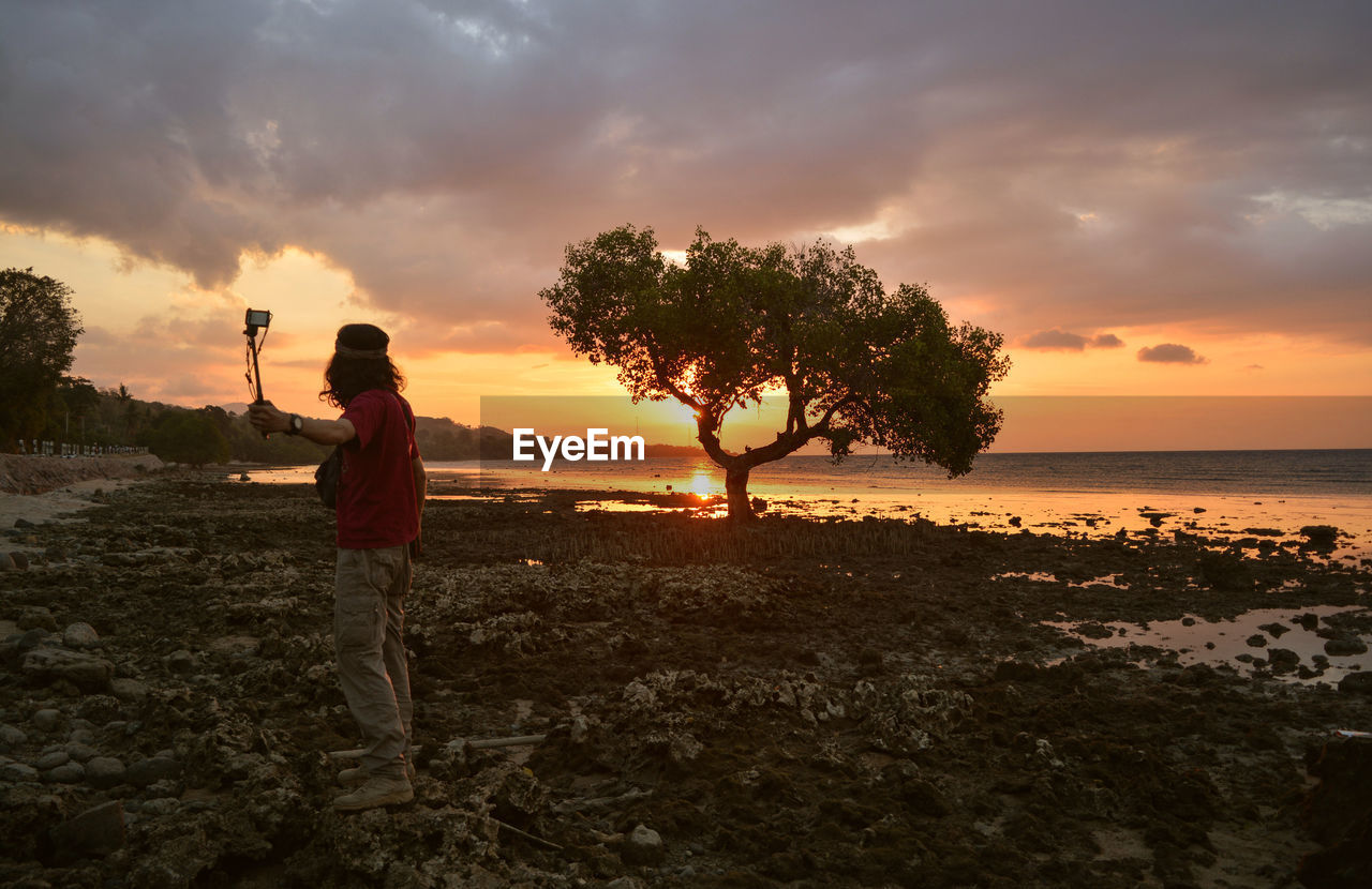 sky, sunset, land, sea, beach, horizon, water, cloud, nature, sunlight, beauty in nature, ocean, dusk, coast, evening, tree, shore, one person, full length, environment, orange color, silhouette, scenics - nature, vacation, holiday, plant, trip, adult, sun, sand, outdoors, leisure activity, landscape, standing, activity, travel destinations, wave, horizon over water, tranquility, travel, men, lifestyles, child, person, childhood, emotion, tropical climate