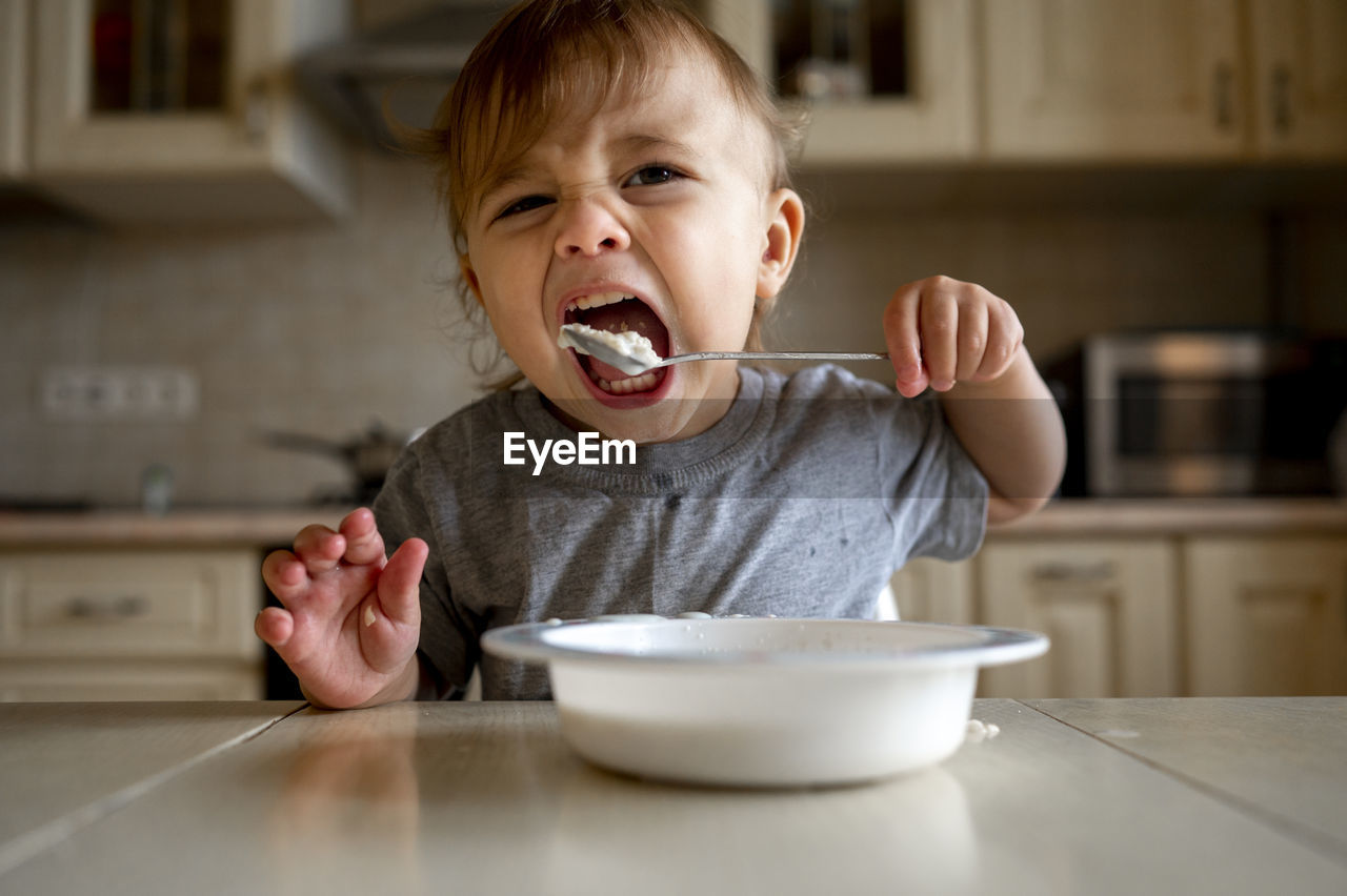 Baby boy eating porridge with spoon in kitchen