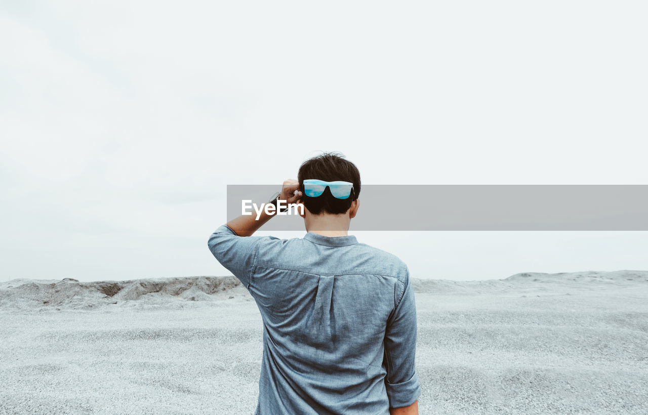 Rear view of man wearing sunglasses standing on beach against sky