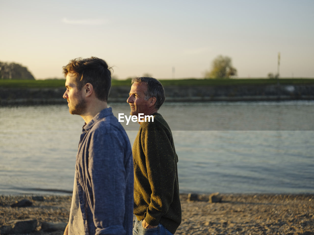 Father and son looking away while spending leisure time by river at evening