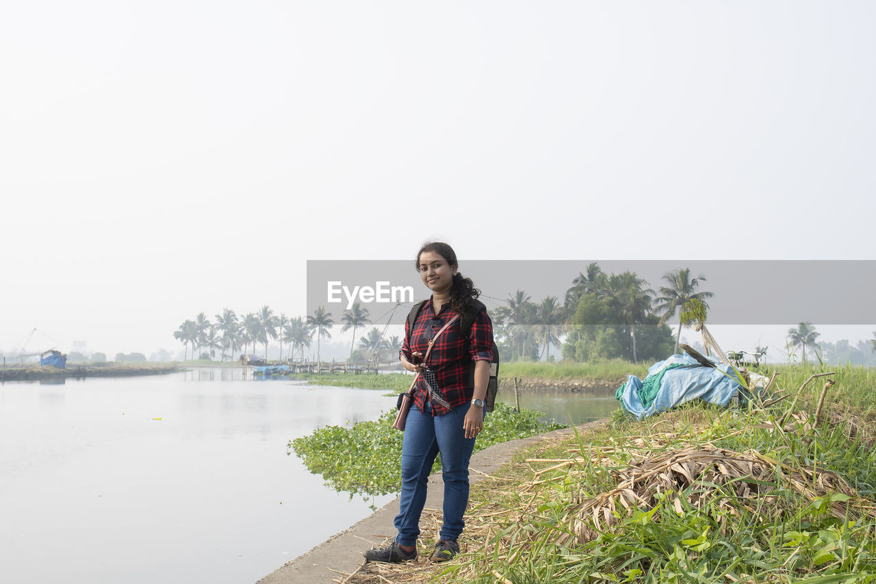 Portrait of young woman standing by river against sky