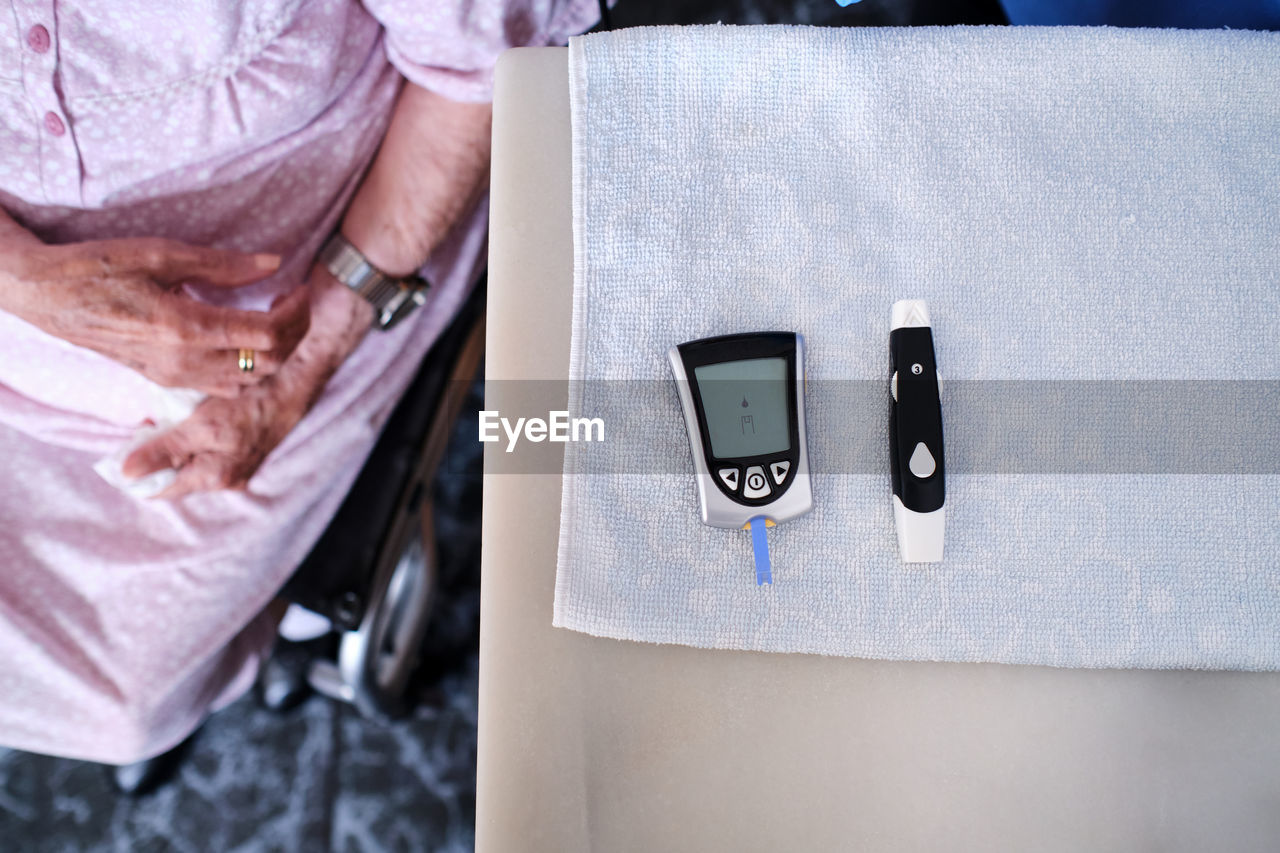 Top view of crop anonymous elderly female sitting in wheelchair near table with special glucose meter in light room at home