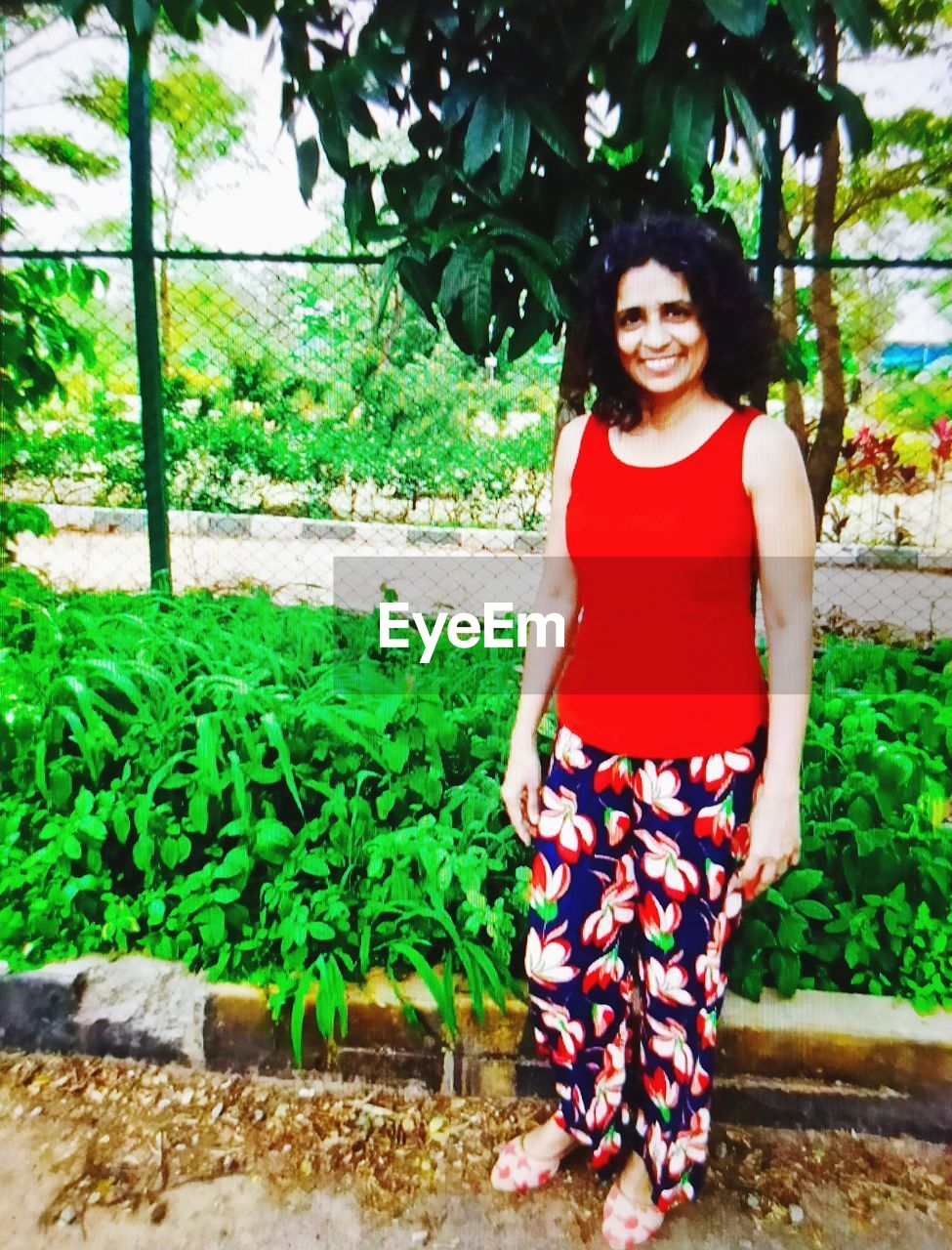 PORTRAIT OF HAPPY YOUNG WOMAN STANDING AGAINST PLANTS