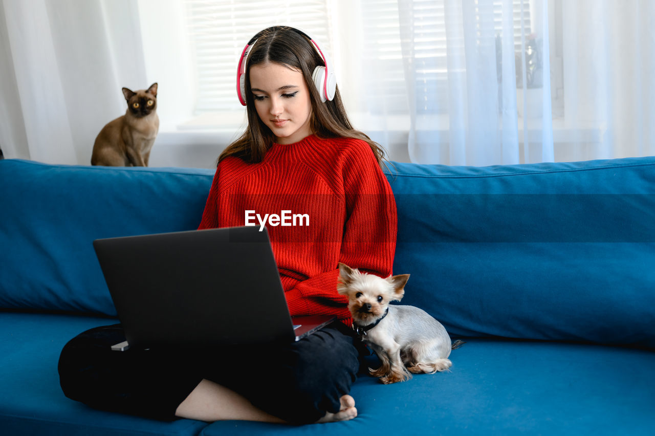 A young girl in a red  sweater works on a laptop at home. her cat and dog are sitting next to her