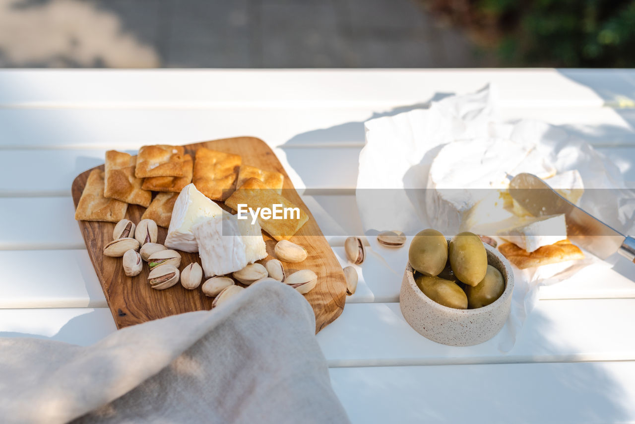 Wine appetizers namely pistachios, camembert cheese, saltine crackers, and olives on a white table.